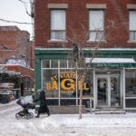 A snowy winter scene of a person with a stroller passing St-Viateur Bagel Shop in Montréal.