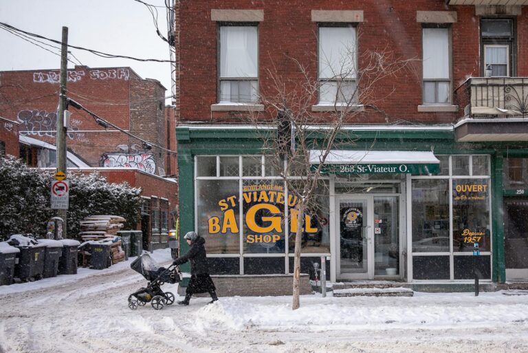 A snowy winter scene of a person with a stroller passing St-Viateur Bagel Shop in Montréal.