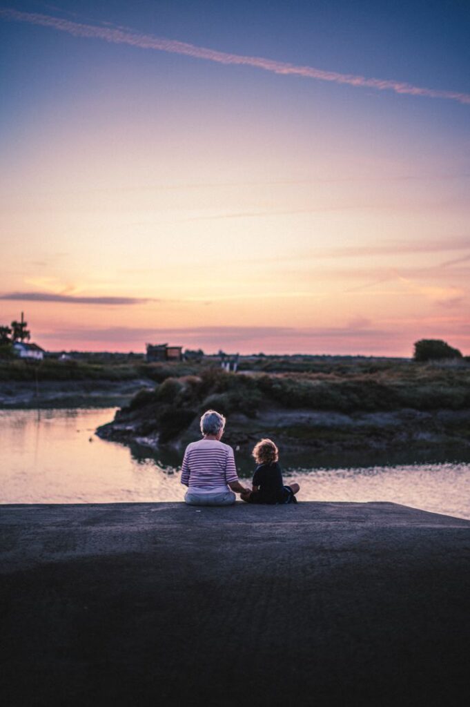 An adult and child sharing a tranquil moment by the water during a vibrant sunset in Arvert, France.